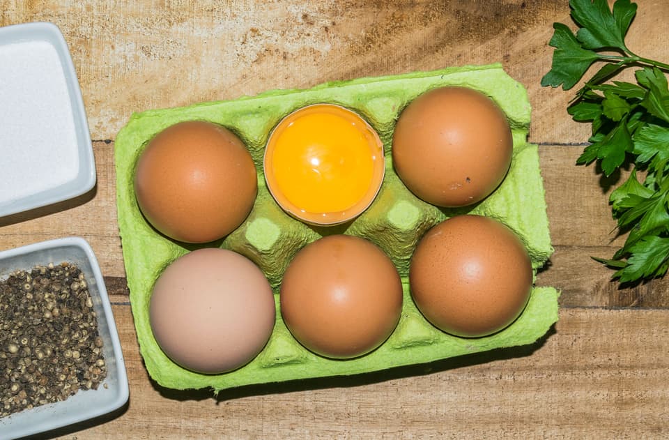 Fresh brown eggs in a carton on a wooden table with parsley and pepper (illustrative)