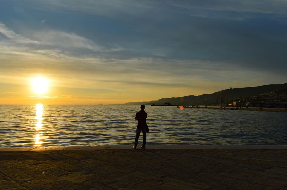 Lone figure seen from a distance standing on the shore as the sun sets over the sea (illustrative)