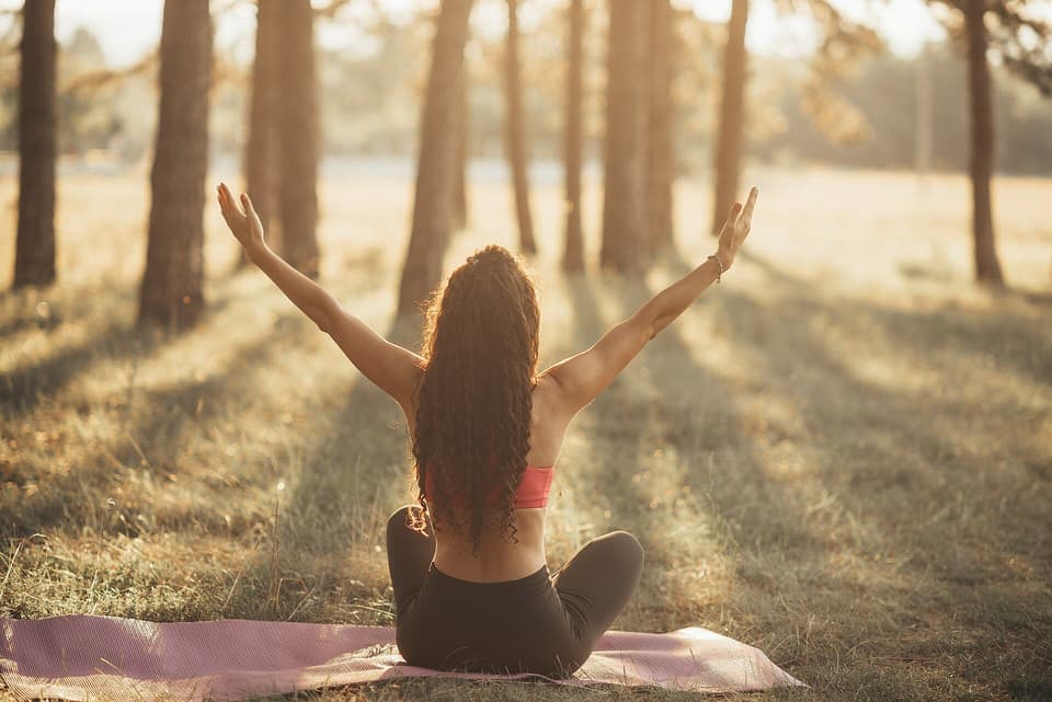 A woman seen from behind doing yoga on a mat among trees in warm light (illustrative)