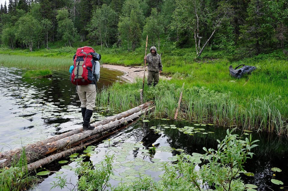 A hiker with a red backpack, seen from behind, crossing a creek on a log (illustrative)